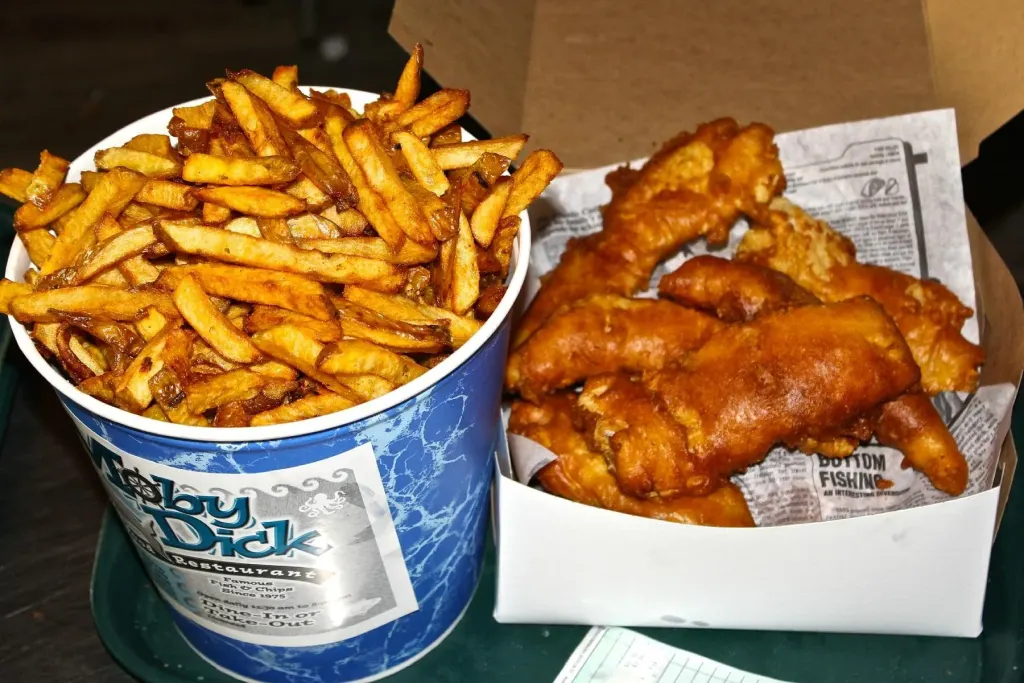 A Moby Dick branded take-out barrel filled with hand-cut fries beside an open box of battered fish pieces on a serving tray.