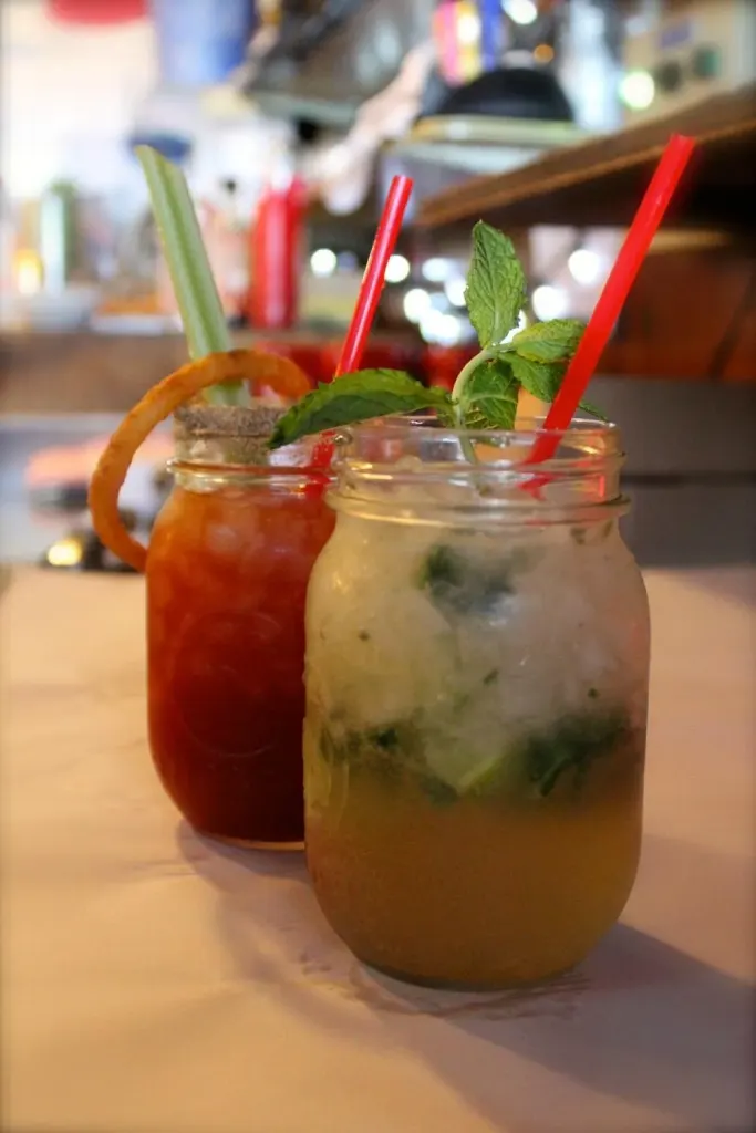 Two cocktails served in mason jars on a table inside the restaurant: a Caesar with a celery stick and a mojito with mint and lime.