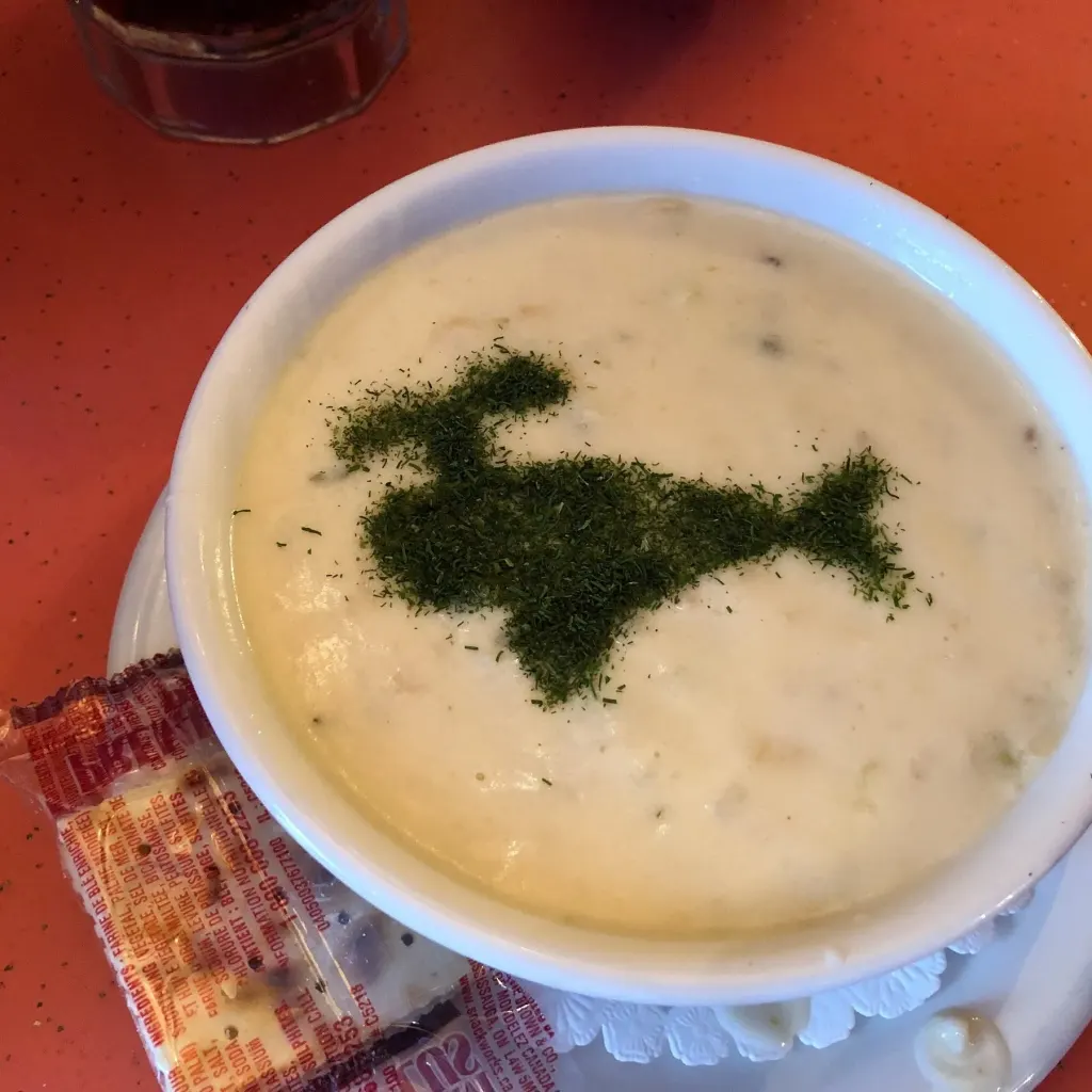 A bowl of creamy clam chowder topped with chopped dill, served on an orange table with a packet of crackers beside it.