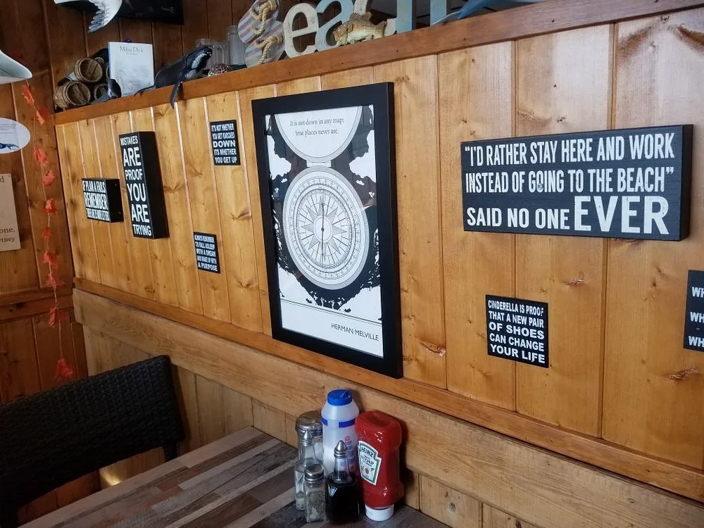 A wood-panelled dining room wall hung with framed sayings, including one reading "I'd rather stay here and work instead of going to the beach, said no one ever", above a booth with ketchup and vinegar.