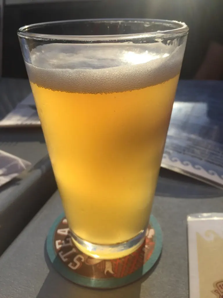 A glass of pale draft beer on a Steamworks coaster, lit by afternoon sun on a patio table.