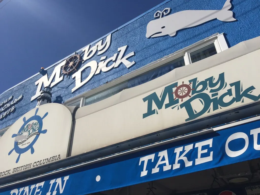 The Moby Dick sign and whale on the blue wall against a clear sky, with the Take Out awning across the bottom of the frame.