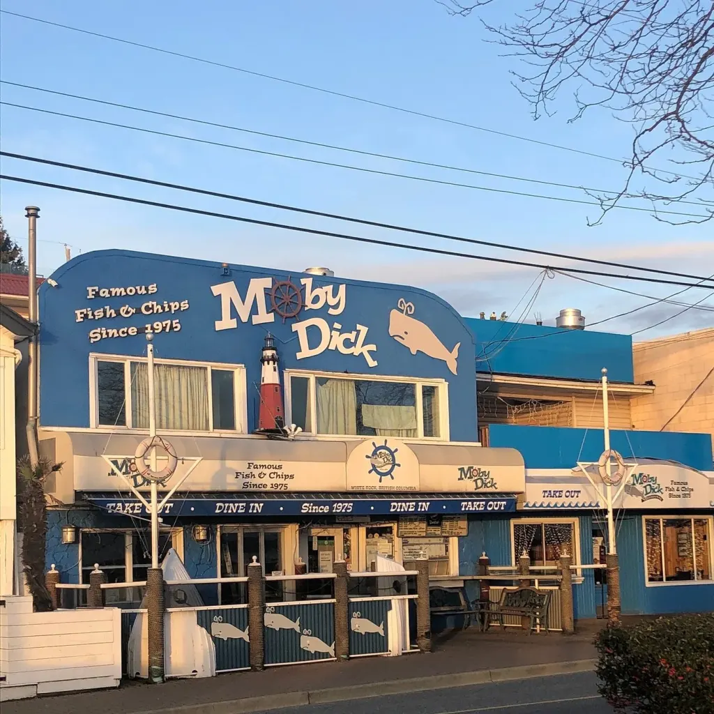The Moby Dick storefront at dusk with the sign lit by the last light, the patio railing in front and the neighbouring buildings behind.