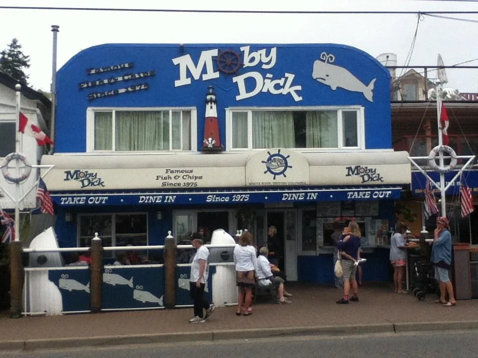 Customers lined up at the take-out window under the blue awning on a sunny afternoon, with the patio and flags to the left.