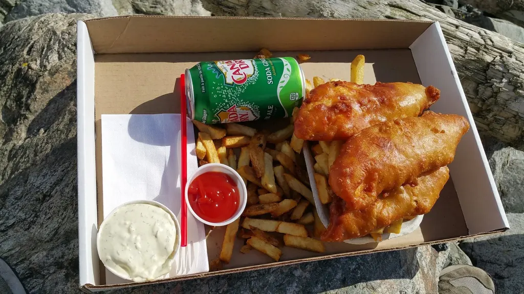A take-out box of battered fish and hand-cut fries with tartar sauce and ketchup, set on the beach rocks with a can of ginger ale beside it.