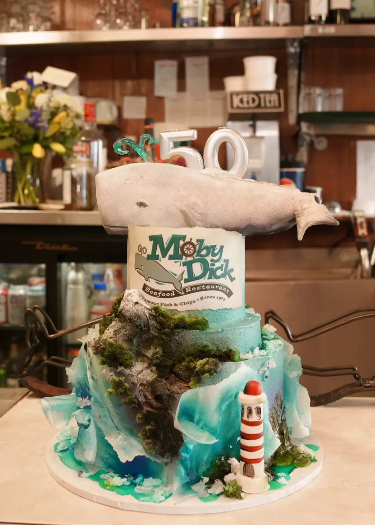A tiered blue cake with ocean waves, a lighthouse and a Moby Dick sign, topped with a white whale and the number 50, on the restaurant counter.