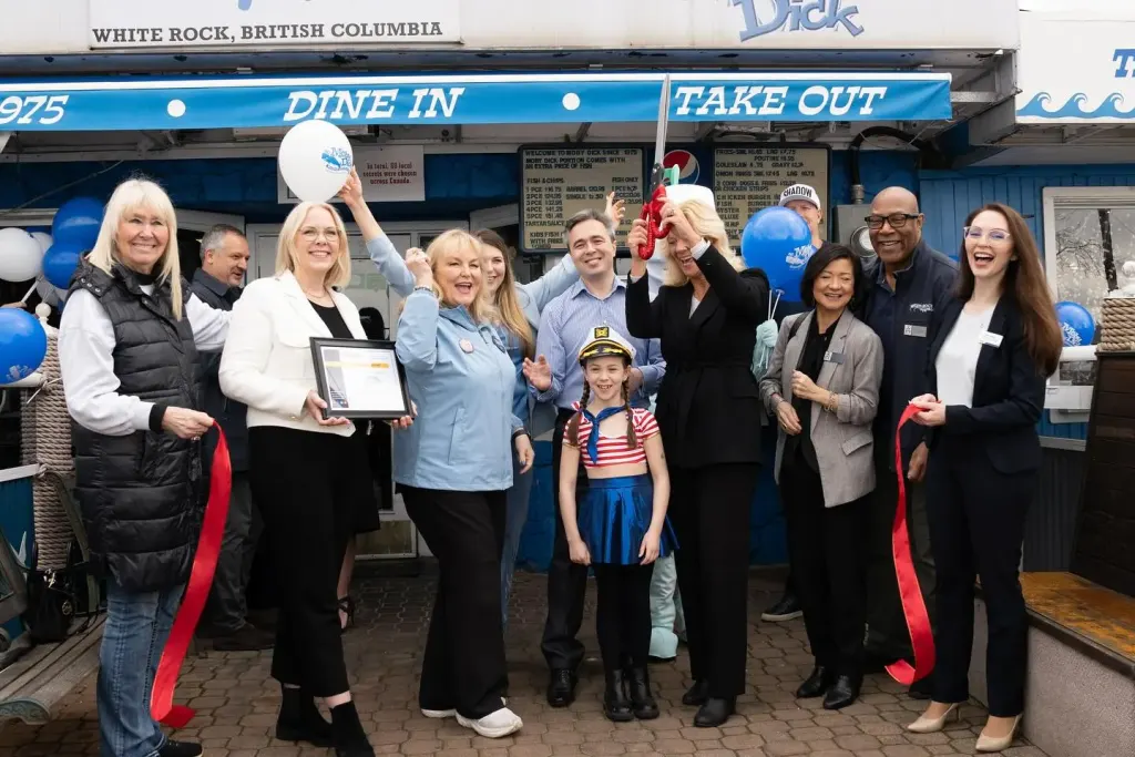 Under the Dine In and Take Out awning, a woman raises a pair of red scissors over the cut ribbon while guests cheer around her, among them a girl in a sailor hat and striped top, and blue and white balloons.