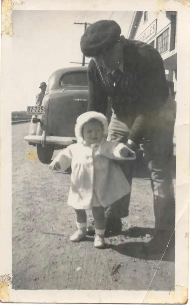 A man in a cap helping a toddler in a white coat walk beside a parked car.