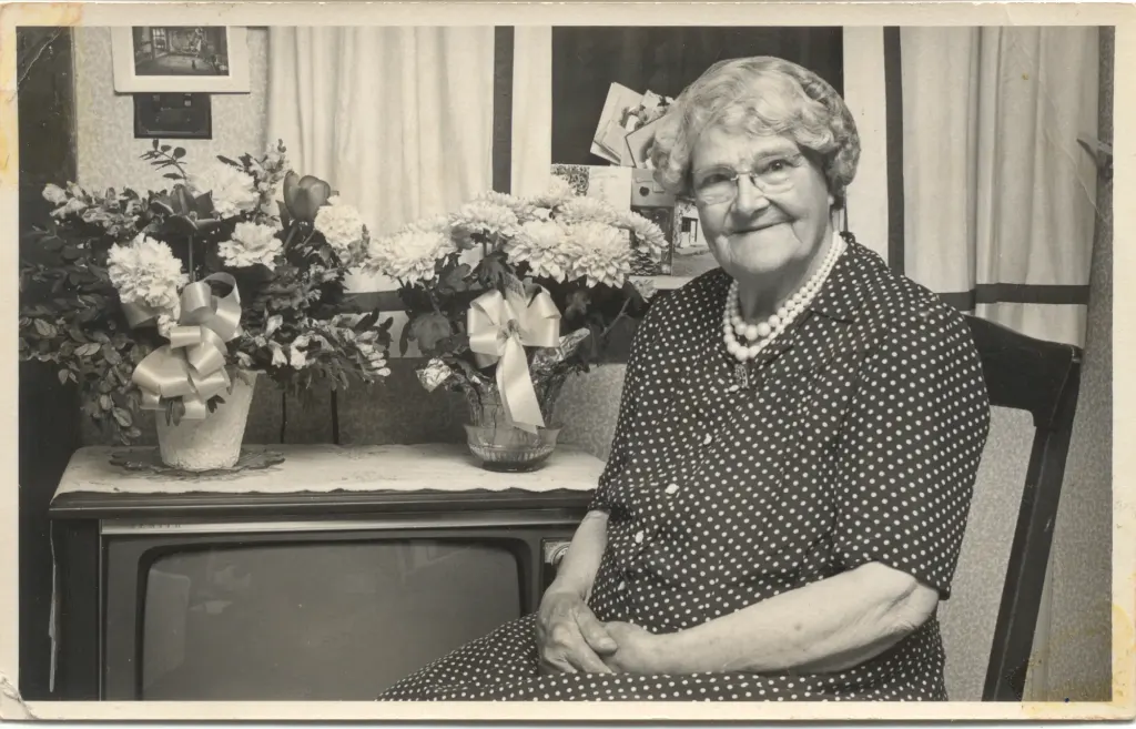 An older woman in a polka-dot dress beside bouquets of flowers set on a television.