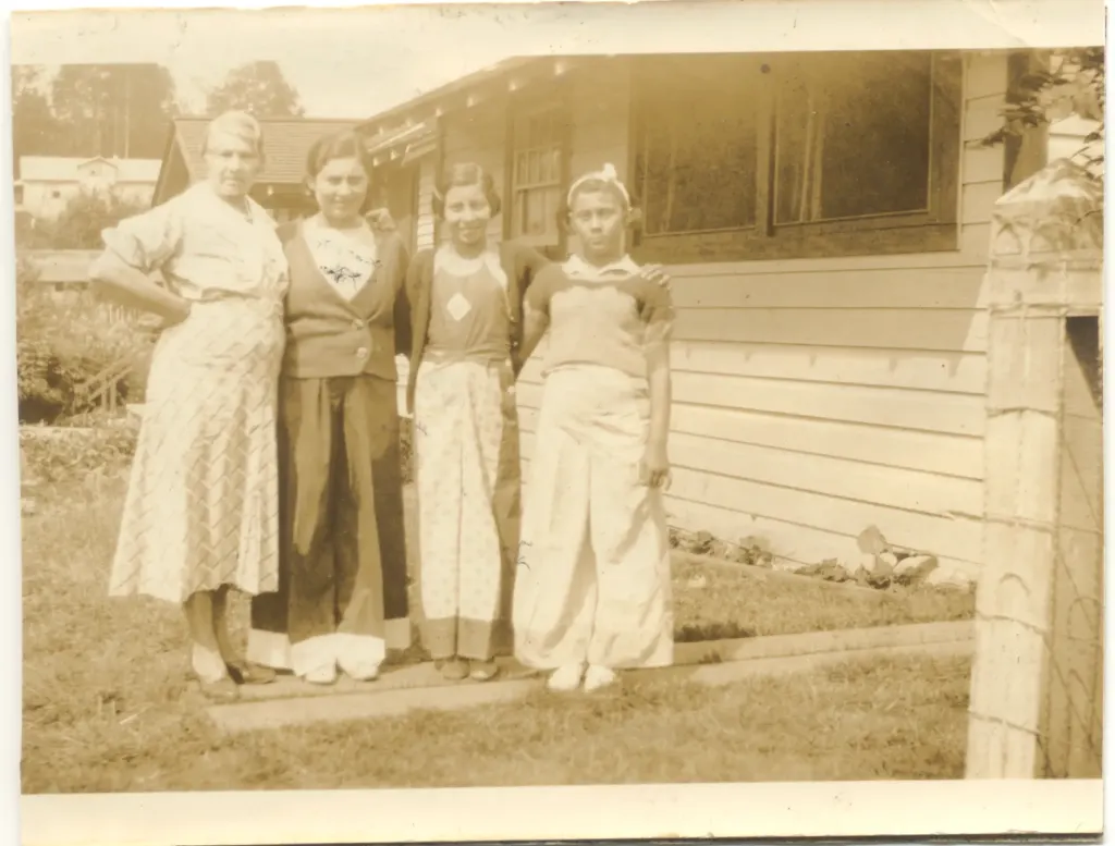 Four women standing on the grass beside a wooden house.