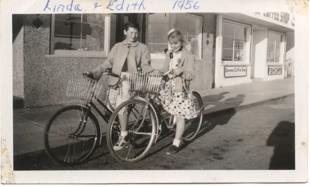Two girls with bicycles on the sidewalk in front of the shops, captioned Linda and Edith 1956 in ink.