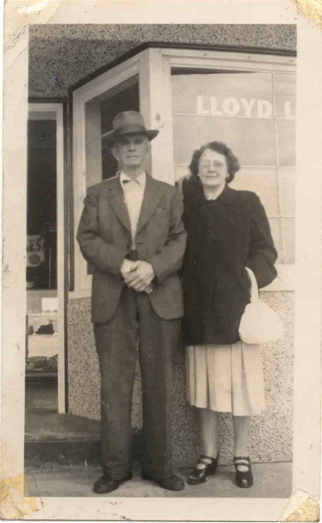 An older man in a hat and an older woman with a white handbag in front of a shop window lettered Lloyd L.