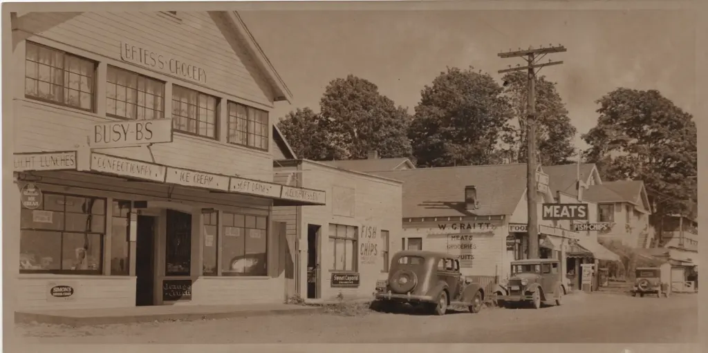 A street of low wooden shops in the late 1920s, with Bates's Grocery on the left and a small fish and chips shop on the right.