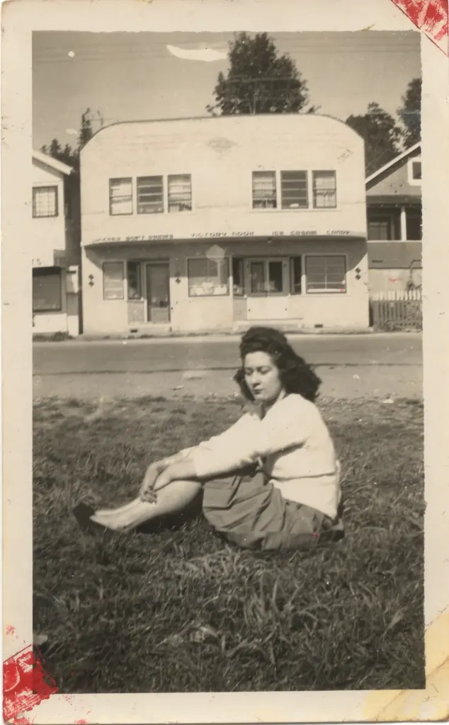A young woman sitting on the grass in front of a two-storey building.