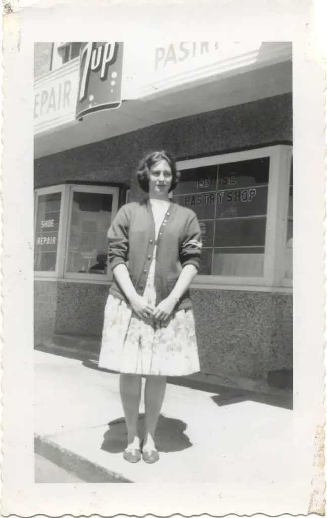 A young woman in a cardigan standing in front of the pastry shop and shoe repair windows, under a 7 Up sign.