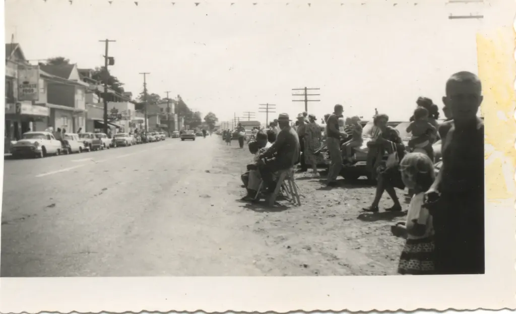 People seated along the curb waiting for a parade, with shops behind.