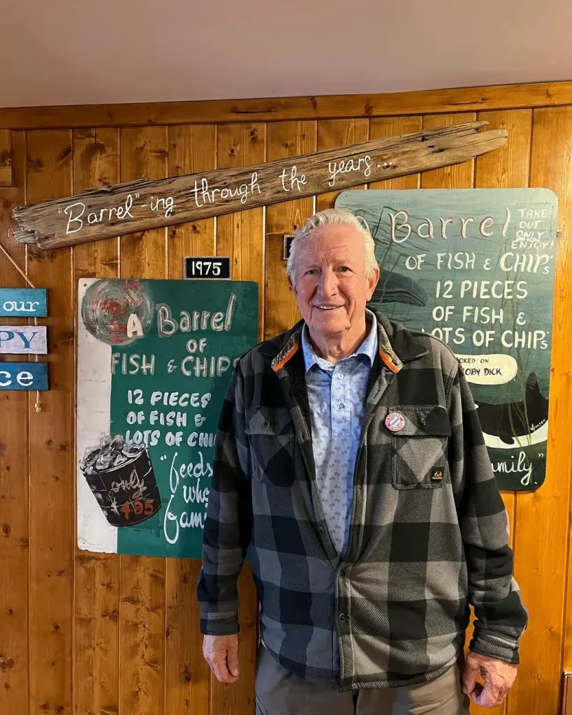 An older man in a plaid jacket smiles in front of wood panelling hung with old signs for a barrel of fish and chips, a 1975 plaque, and a driftwood sign reading Barrel-ing through the years.