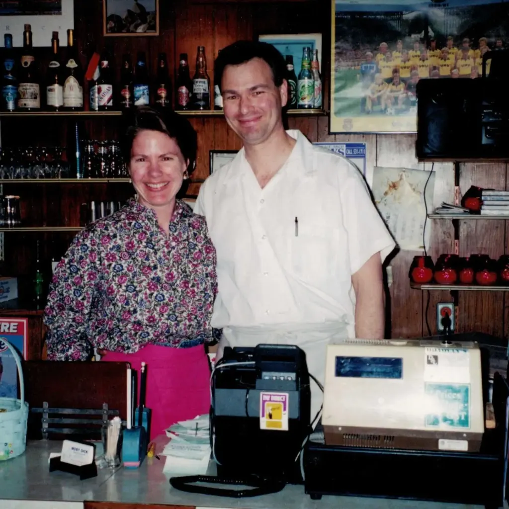 A smiling couple behind the counter, with bottles on the shelves and framed photographs on the wall behind them.
