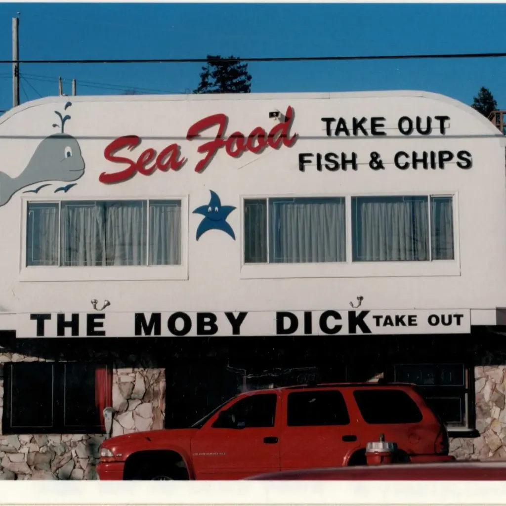 The old storefront with a whale and "Sea Food, Take Out, Fish & Chips" painted on the sign, "The Moby Dick" along the awning, and a red car parked in front.