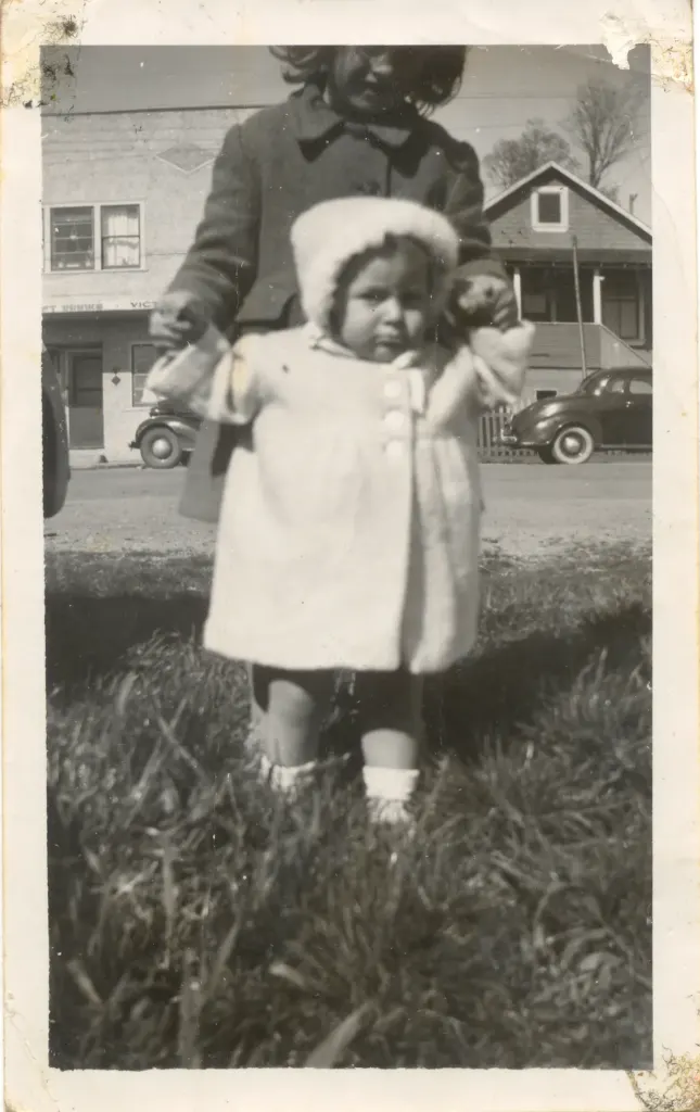 A toddler in a white coat and bonnet standing on the grass, with houses and a car behind.