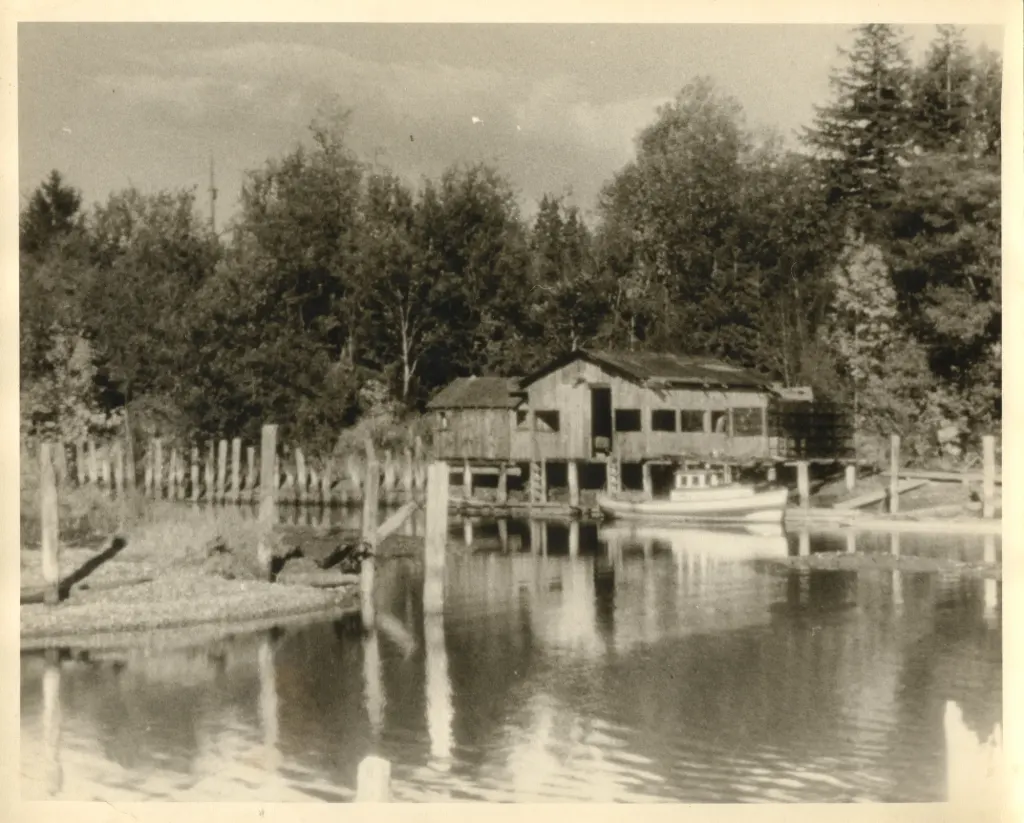 A wooden boathouse on pilings beside still water, with a boat moored and tall trees behind.